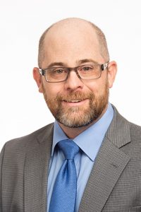 Professional headshot of a man with a beard, wearing glasses, a gray suit, and a light blue shirt with a blue tie, smiling against a white background. Ideal for business or corporate profiles.